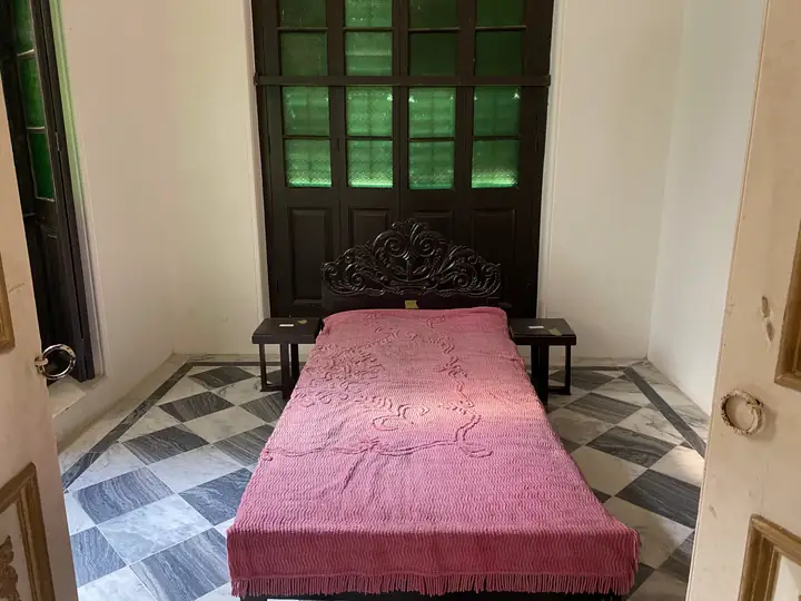 Kamar mungil dengan ranjang bersprei merah muda, lantai tegel kotak-kotak, dan jendela kaca hijau