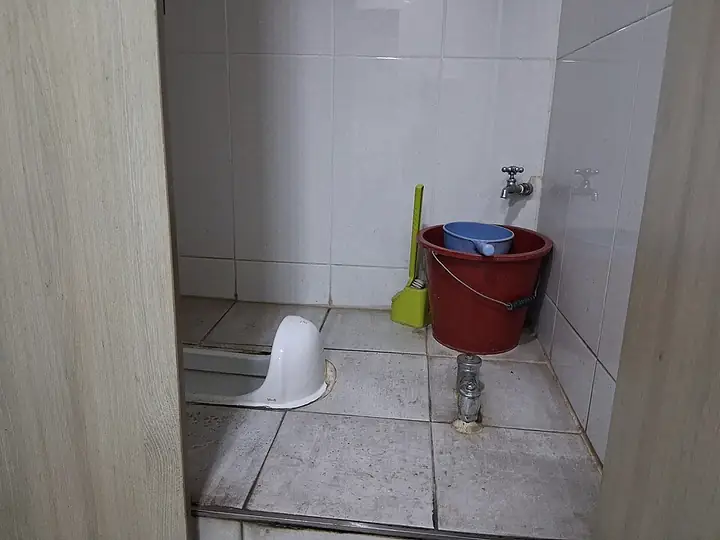 Kamar mandi luar dengan kloset jongkok, ember merah, dan keran